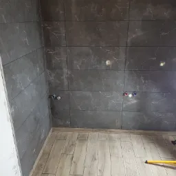 Ciemnoszare płytki imitujące marmur na ścianie łazienki, z widocznymi przyłączami wodnymi i fragmentem oświetlenia budowlanego oraz butelką z rozpylaczem na podłodze z paneli.