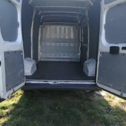 Wnętrze pustego, białego busa z otwartymi tylnymi drzwiami, widoczne na tle trawnika w słoneczny dzień.