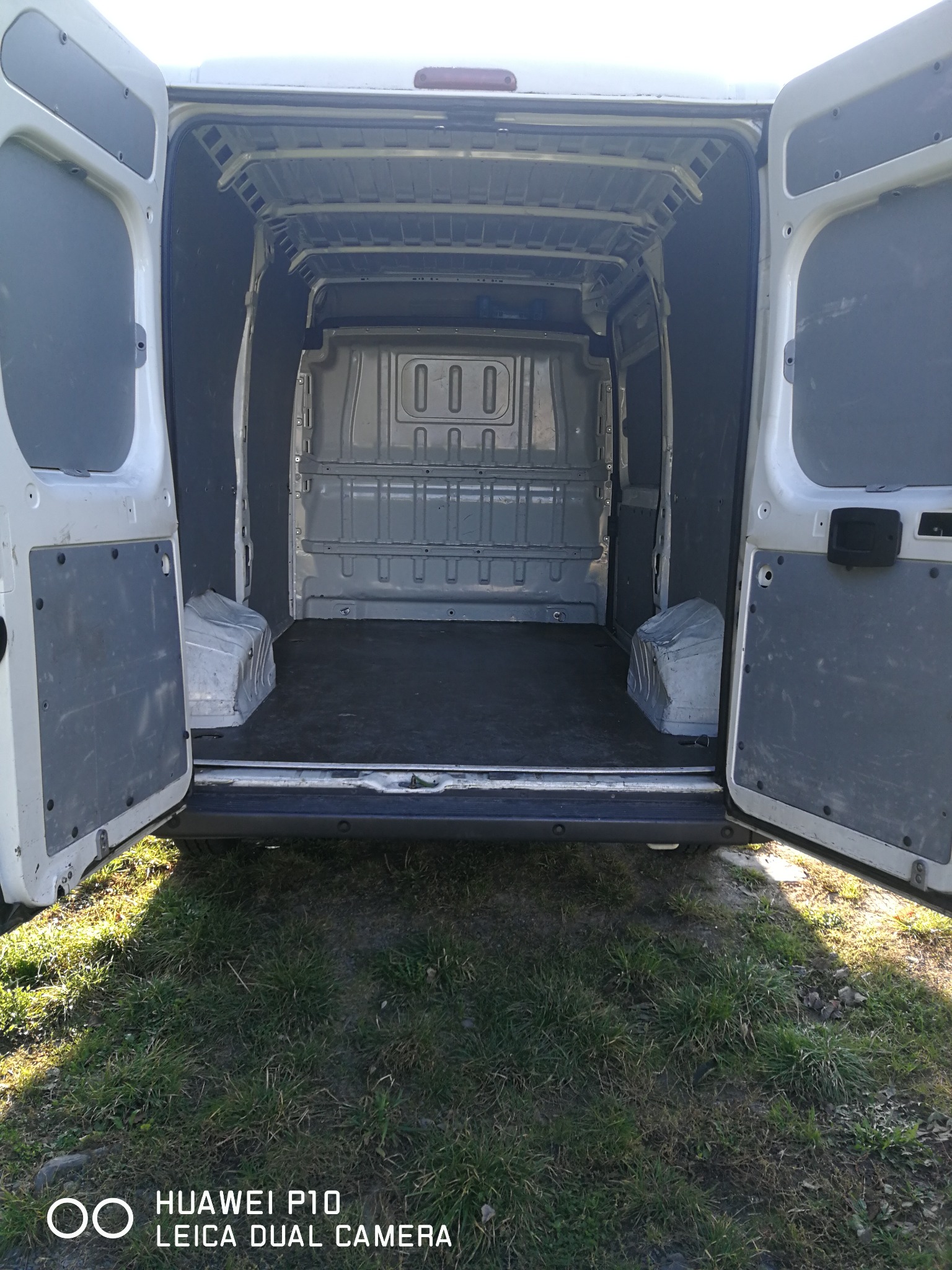 Wnętrze pustego, białego busa z otwartymi tylnymi drzwiami, widoczne na tle trawnika w słoneczny dzień.