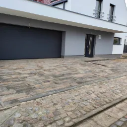 Nowoczesny podjazd z kostki brukowej w odcieniach szarości i brązu przed domem z szarą bramą garażową, widoczna skrzynka elektryczna i fragment chodnika z kostki granitowej.