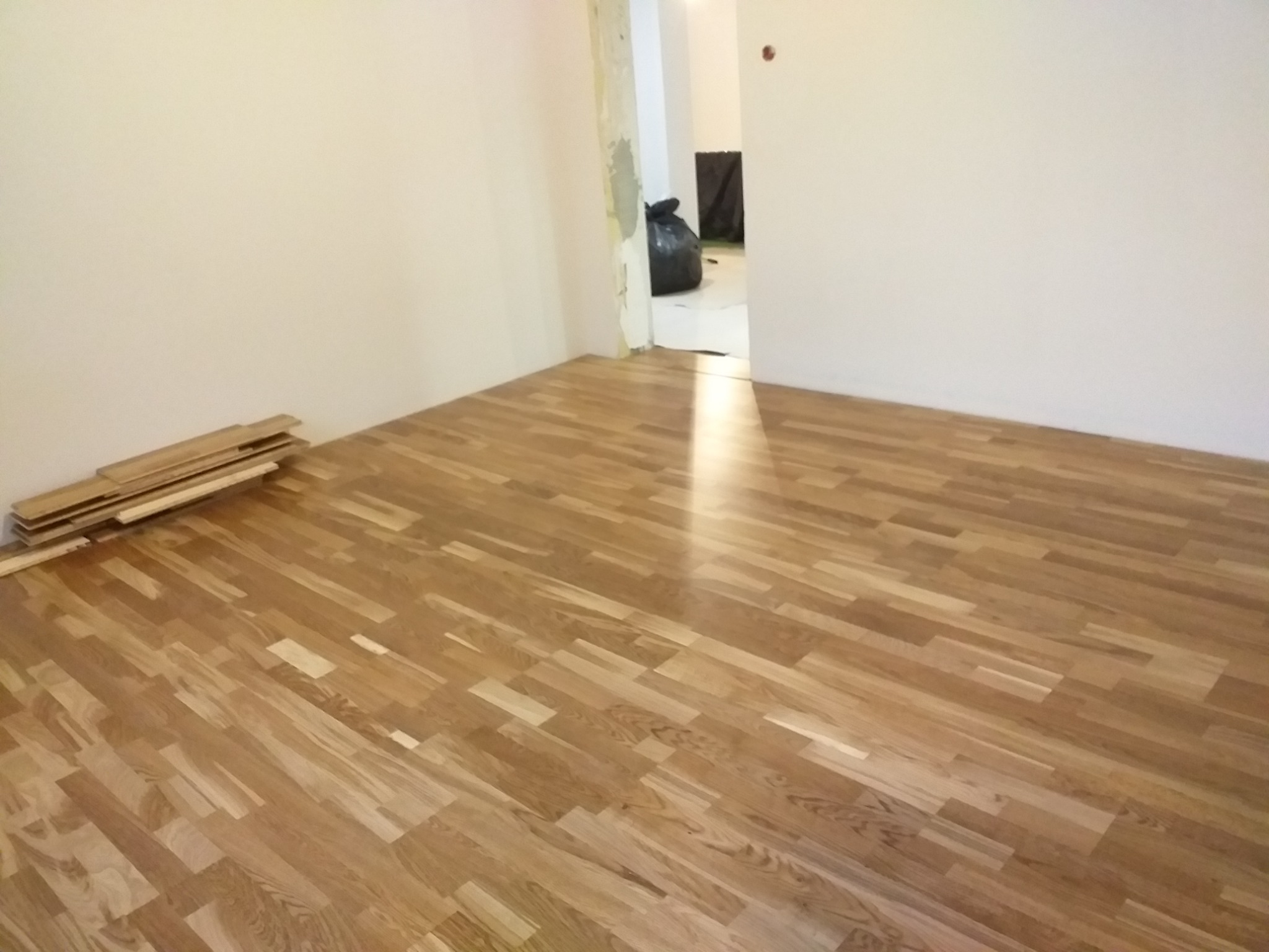 Fragment pokoju z ułożonym parkietem w odcieniach brązu, z paczką desek leżącą przy ścianie i workiem na śmieci widocznym w przejściu do innego pomieszczenia.