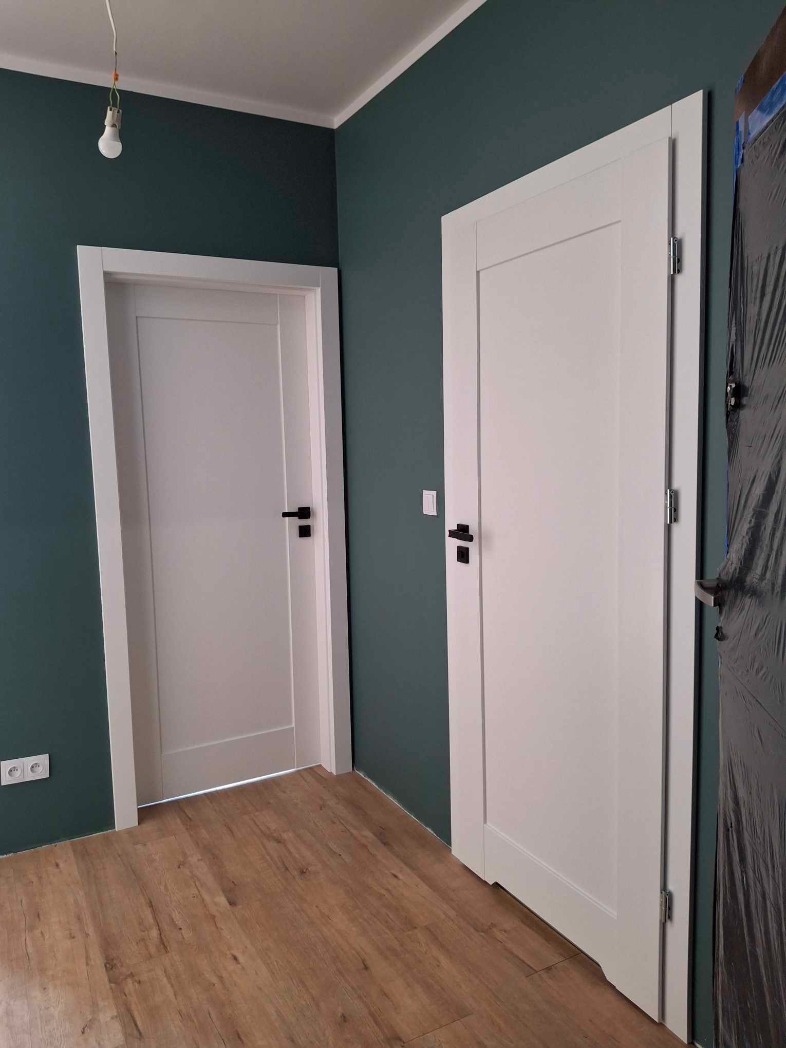 Dwa białe, wewnętrzne drzwi z czarnymi klamkami na tle ciemnozielonej ściany i drewnianej podłogi; jeden drzwi zabezpieczony folią malarską.