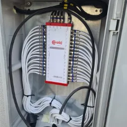 Rozdzielacz antenowy marki Crab w skrzynce z licznymi białymi kablami koncentrycznymi i czarnymi przewodami, widoczne złącza F i zasilacz.