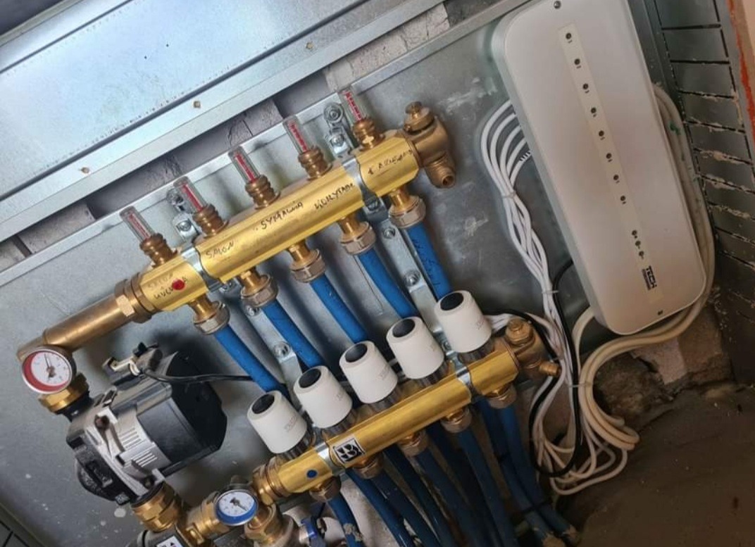 Rozdzielacz podłogowy z pompą obiegową i sterownikiem, widoczne rury doprowadzające wodę, zamontowany w szarej skrzynce, z napisami odręcznymi na mosiężnych elementach.