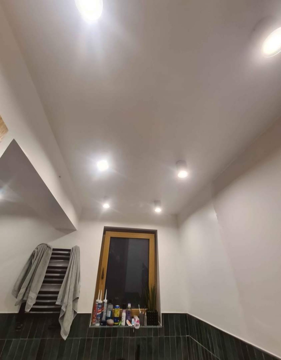 Łazienka po remoncie z widocznymi punktowymi lampami sufitowymi, grzejnikiem drabinkowym z suszącym się ręcznikiem i oknem z rośliną doniczkową oraz kosmetykami na parapecie.