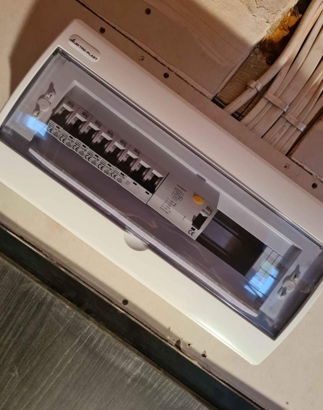 Zamknięta skrzynka bezpiecznikowa marki Elektro-Plast z widocznymi bezpiecznikami IDEAL, zamontowana na ścianie z wystającymi kablami elektrycznymi.