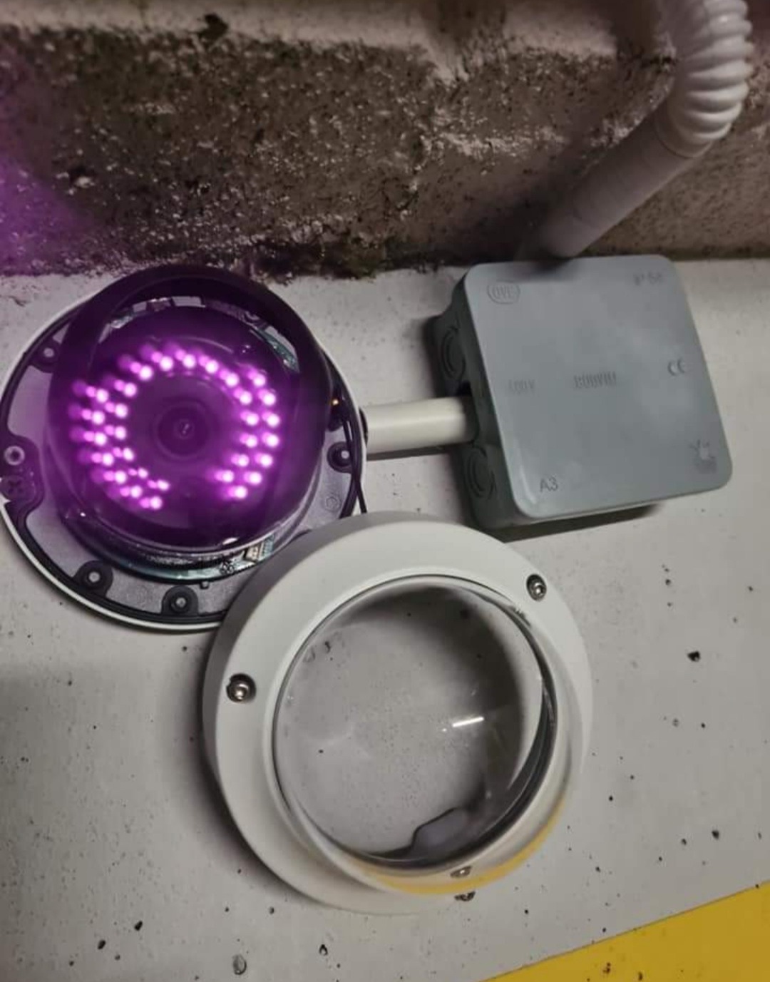 Otwarta kamera monitoringu z włączonymi diodami IR, widoczna puszka instalacyjna i przewód na szarej ścianie.