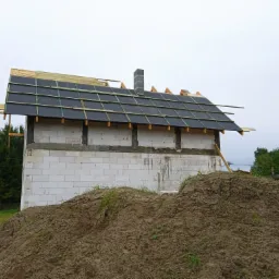 Surowy stan budowy domu: ściany z białego bloczka, częściowo pokryty dach z czarną papą i drewnianą konstrukcją, widoczny komin z szarej cegły oraz hałda ziemi przed budynkiem.
