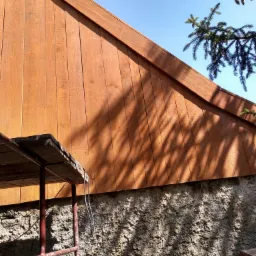 Drewniana elewacja szczytowa domu w trakcie malowania, widoczne rusztowanie, ściana fundamentowa z kamienia i cień drzewa na elewacji.