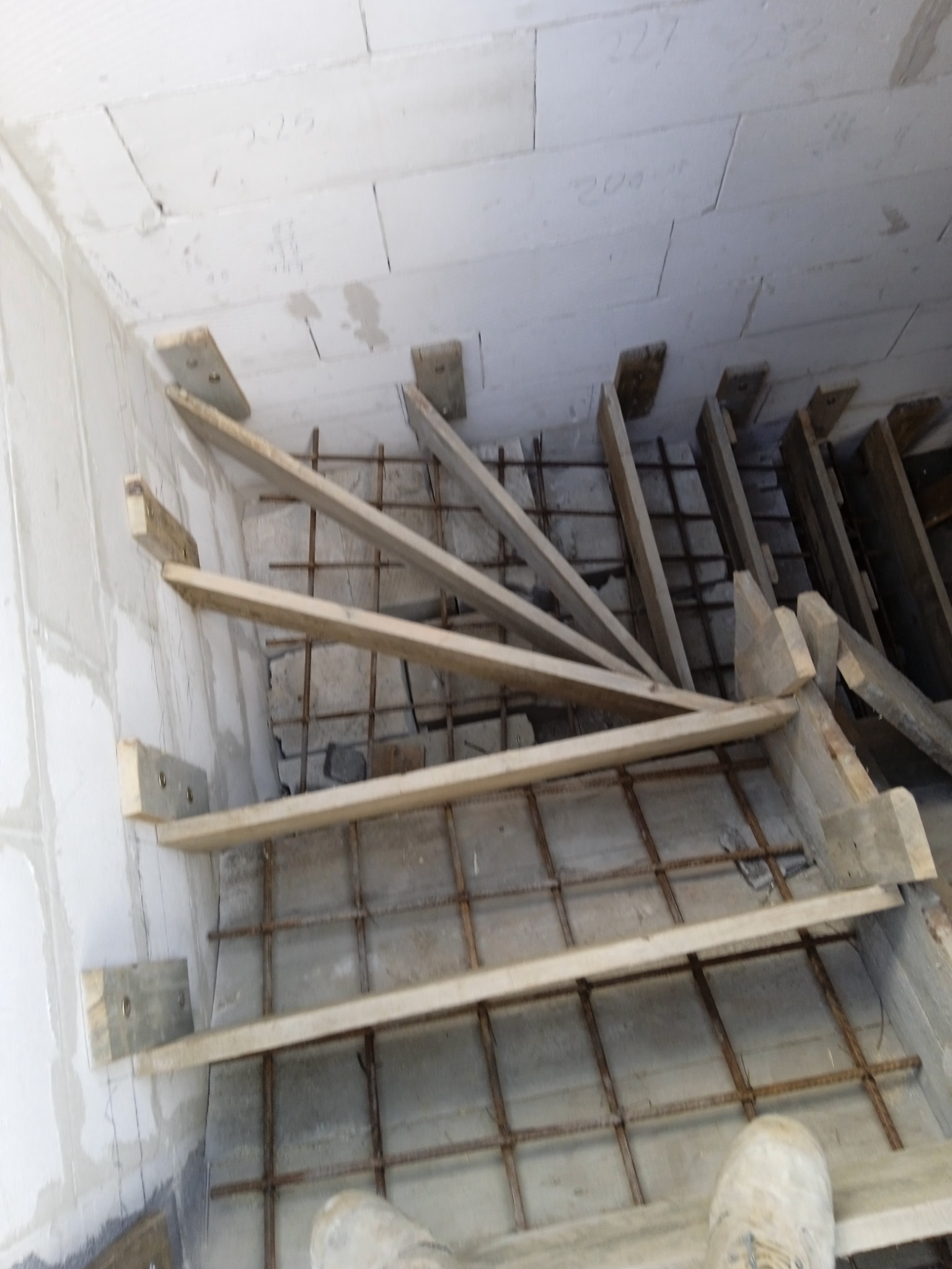 Wewnętrzna konstrukcja schodów żelbetowych z drewnianym szalunkiem i siatką zbrojeniową, widok z góry na buty robotnika.