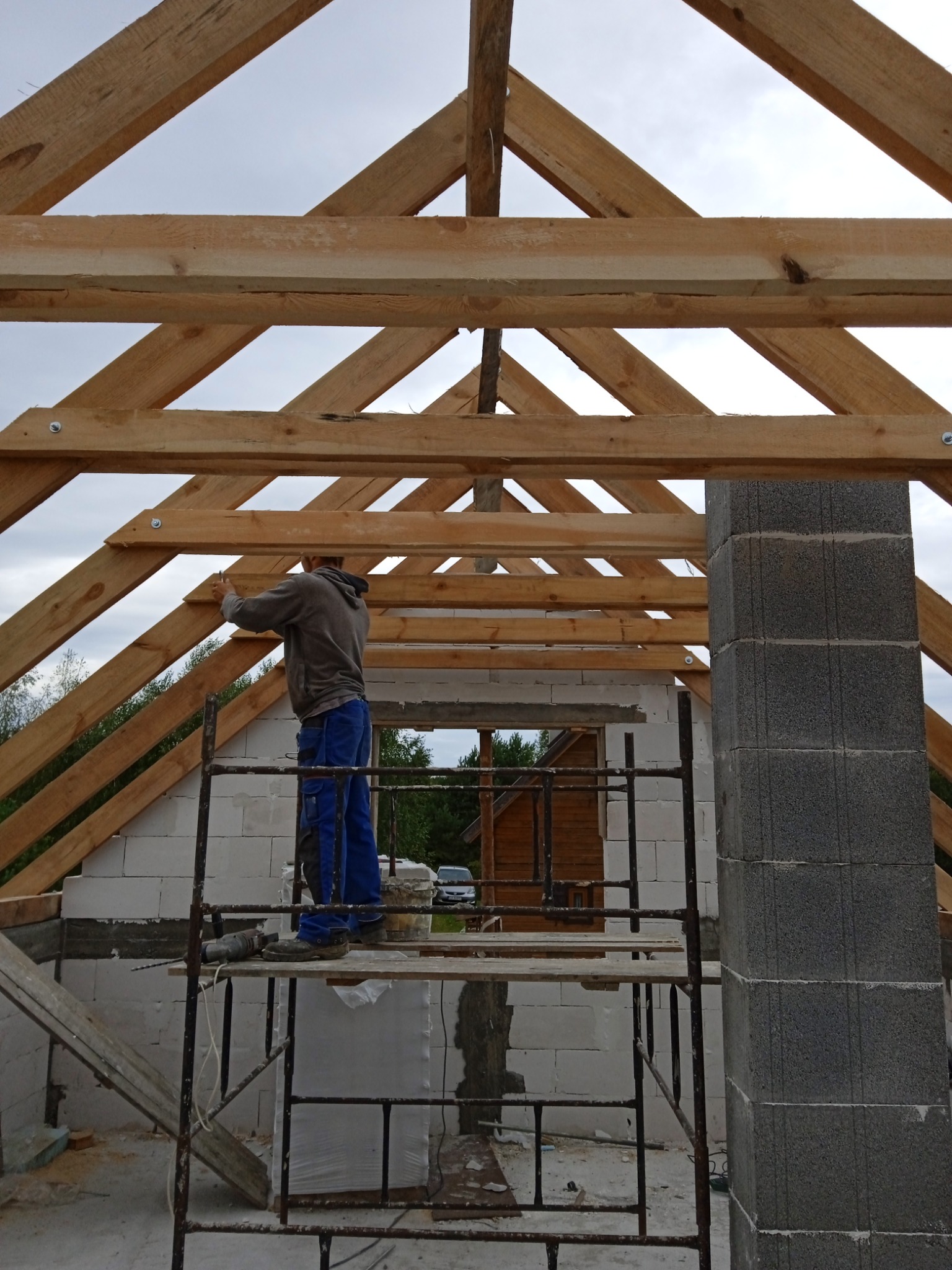 Konstrukcja drewniana dachu w trakcie montażu, widok z wnętrza budynku w stanie surowym, pracownik na rusztowaniu mocuje elementy więźby dachowej.