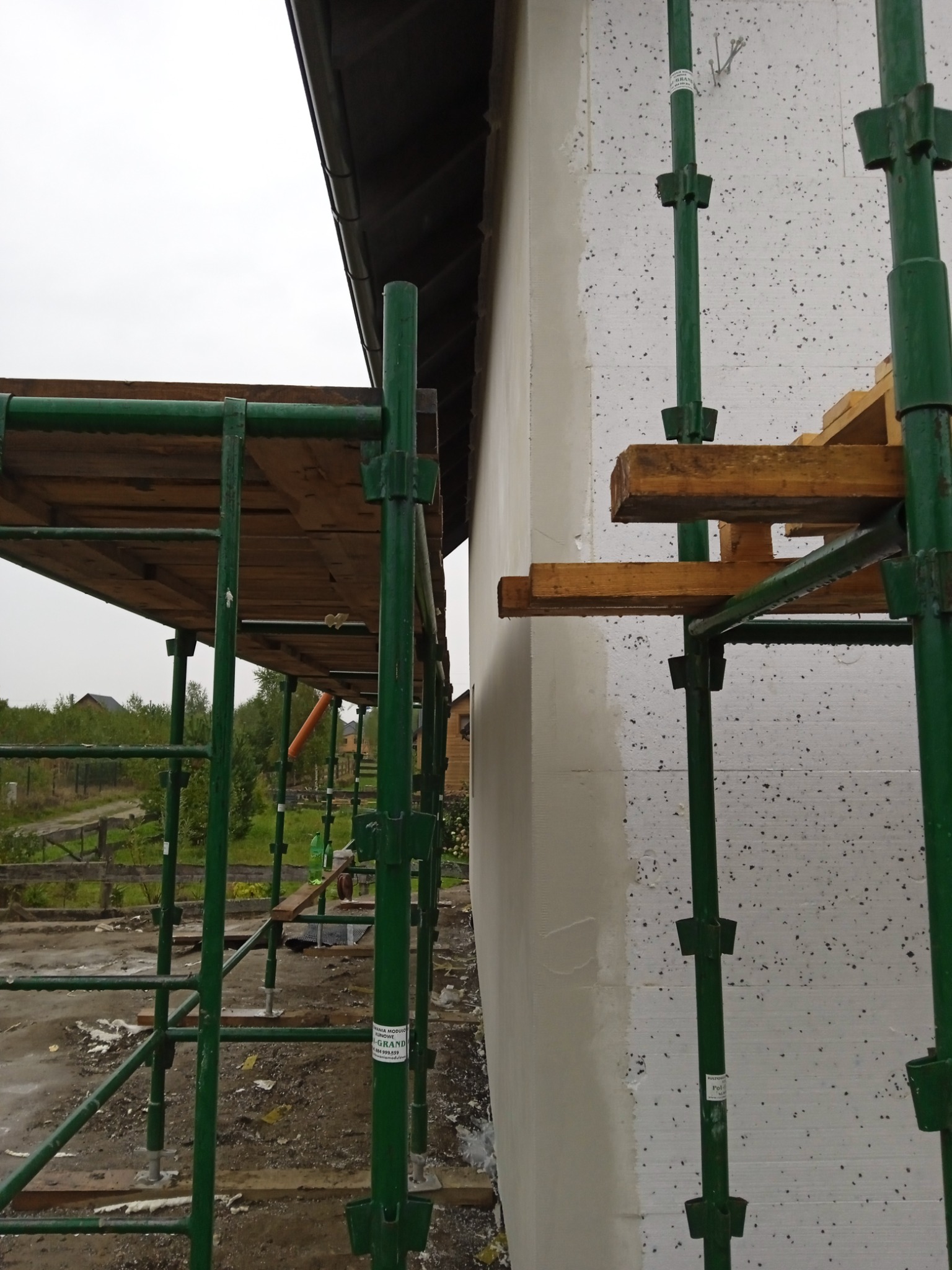 Ściana budynku w trakcie ocieplania styropianem, widoczne rusztowanie z zielonych rur, częściowo otynkowana powierzchnia i elementy drewniane.