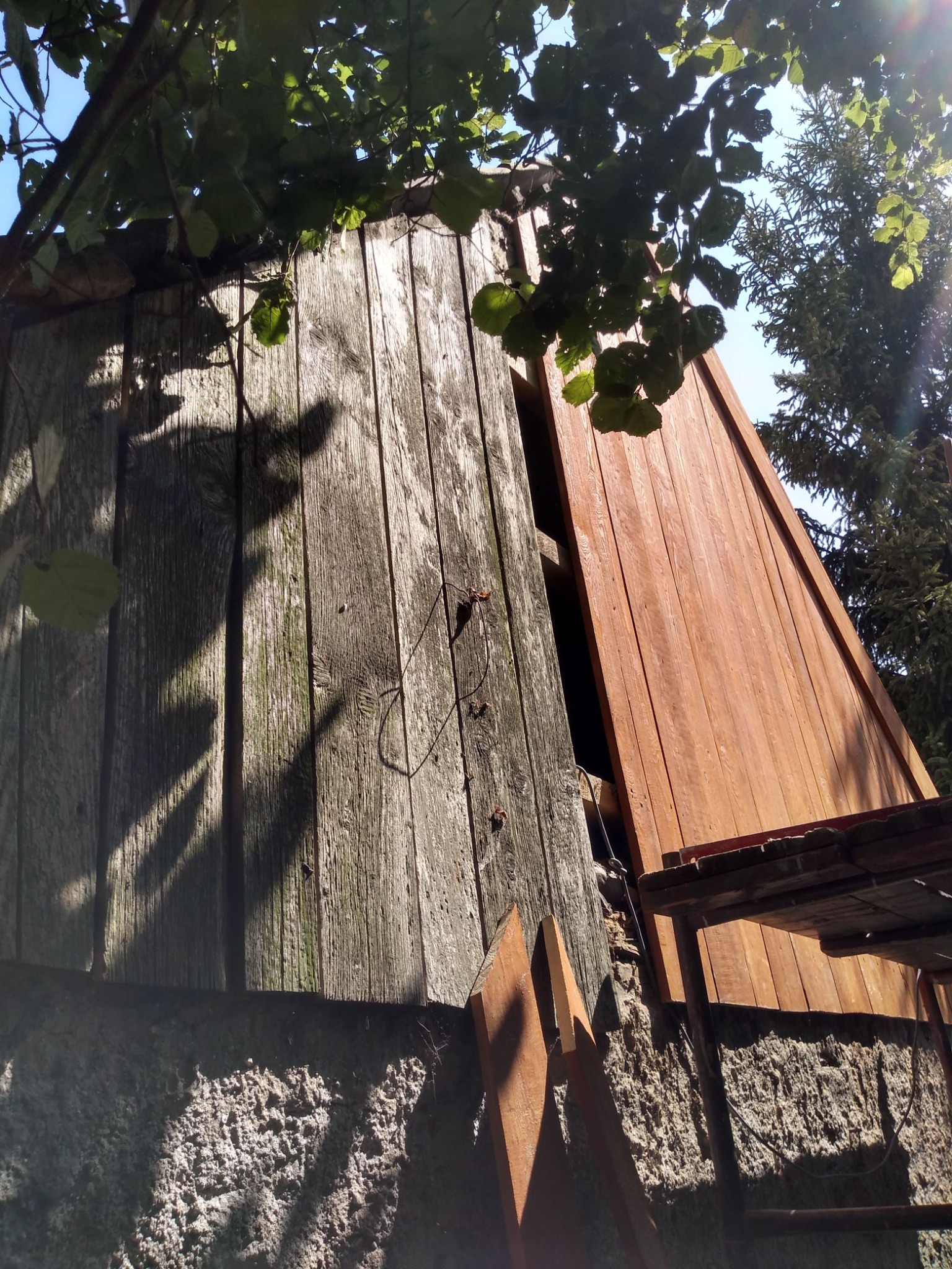 Fragment elewacji budynku w trakcie prac, widoczne deski w różnym stanie: szare, zwietrzałe drewno obok świeżo pomalowanych, brązowych desek. U góry kadru gałęzie drzew i fragment nieba.