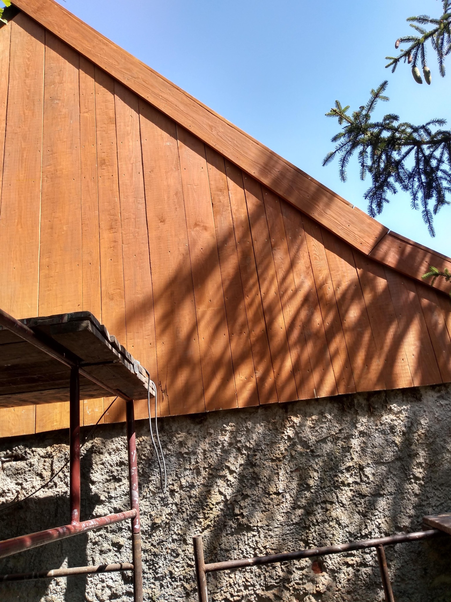 Drewniana elewacja szczytowa domu w trakcie malowania, widoczne rusztowanie, ściana fundamentowa z kamienia i cień drzewa na elewacji.