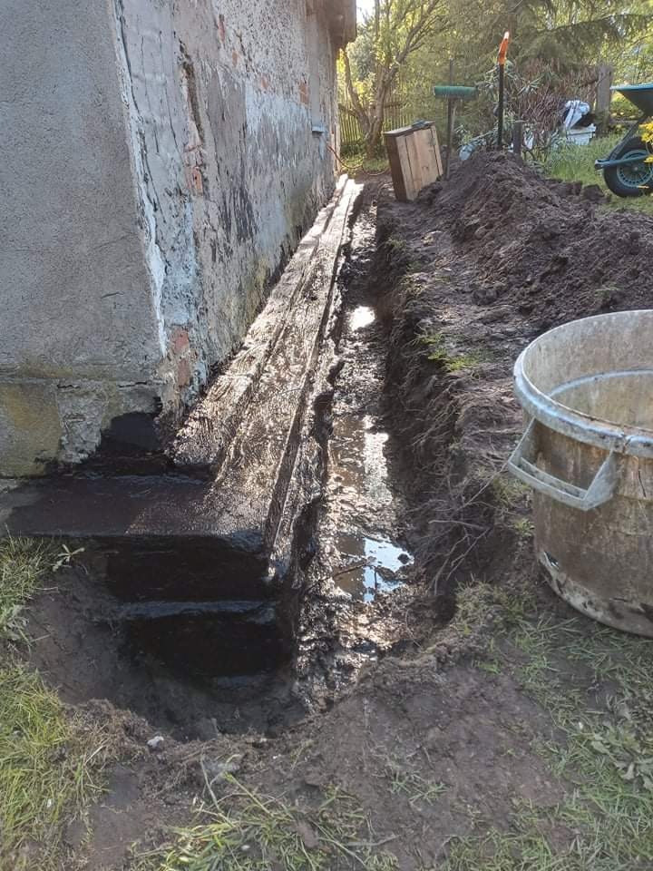 Wykopany rów wzdłuż fundamentu budynku, zabezpieczony drewnianą szalówką, częściowo wypełniony wodą, z widocznym starym murem budynku i narzędziami w tle.