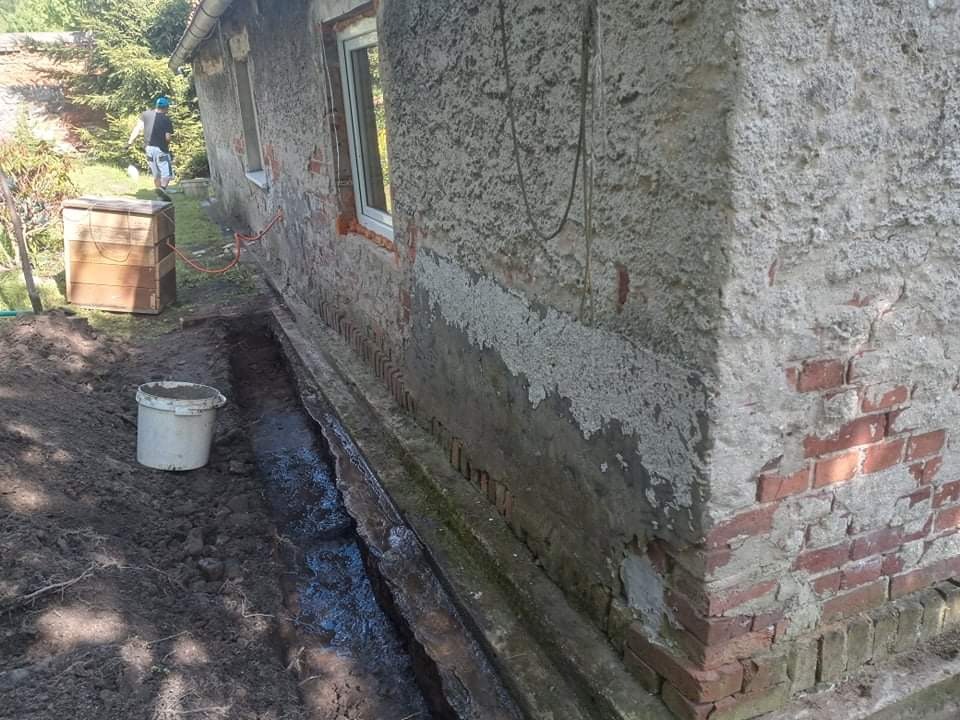 Przygotowanie do prac ziemnych przy starej, częściowo otynkowanej ścianie budynku z odsłoniętą cegłą, widoczny wykop wypełniony wodą i wiadro z zaprawą. W tle osoba w niebieskiej czapce.