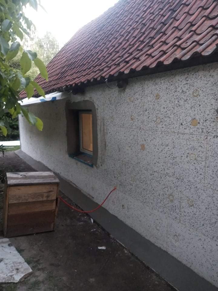 Ściana budynku w trakcie prac elewacyjnych: widoczne warstwy ocieplenia z okrągłymi zaślepkami, świeżo wylany fundament przy ścianie oraz wystający czerwony kabel.