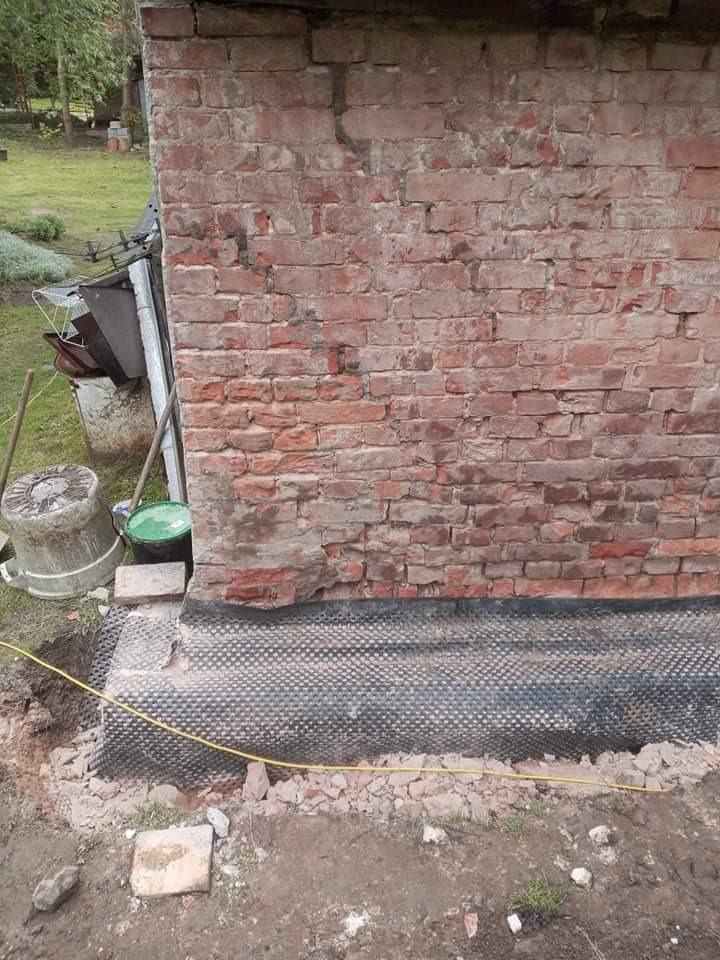 Fragment starej ceglanej ściany z widocznym uszkodzeniem i nową hydroizolacją przy fundamencie, otoczony wykopem ziemnym i gruzem.
