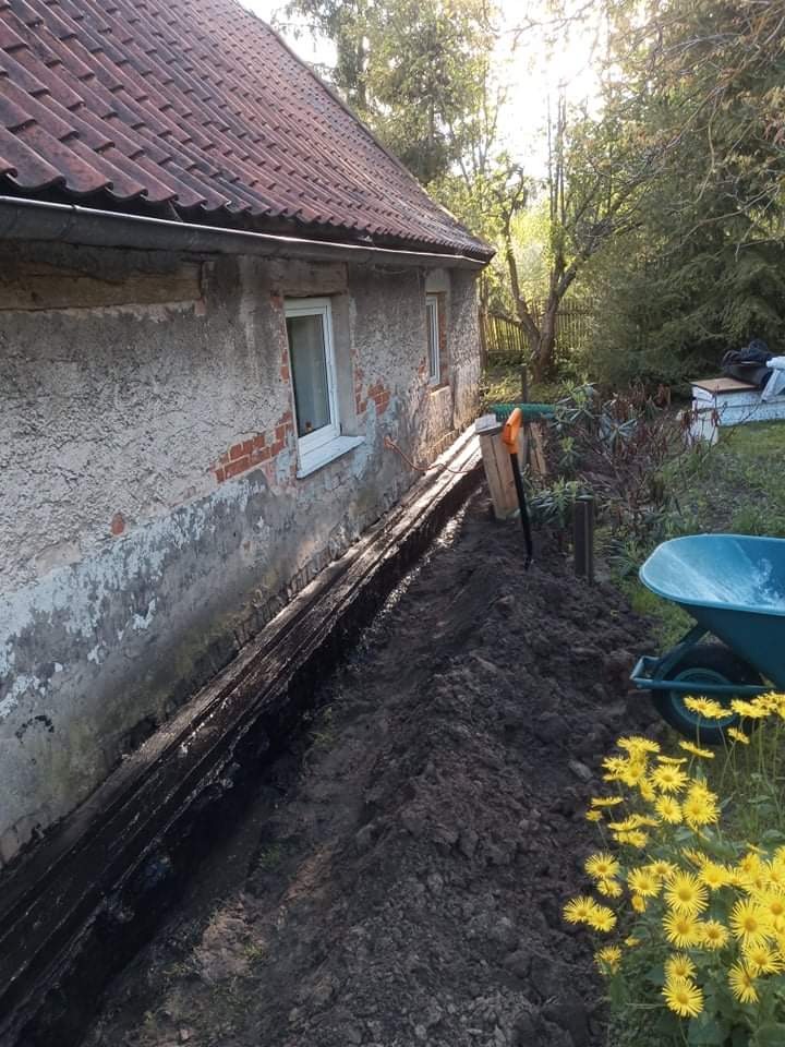 Przygotowanie podłoża pod elewację starego domu: wykopany rów, zabezpieczona ściana, taczka i kwitnące żółte kwiaty w tle.