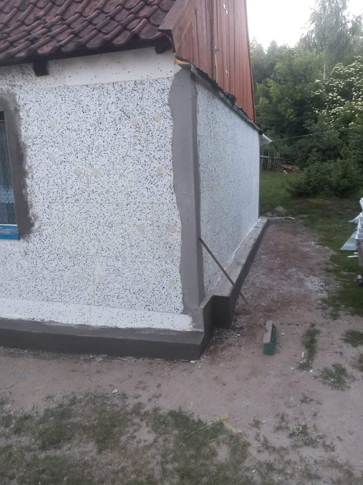 Fragment ściany budynku z widocznym ociepleniem i świeżo nałożonym tynkiem dekoracyjnym typu baranek, z obróbką okienną w trakcie wykonywania, na tle trawnika i drzew.