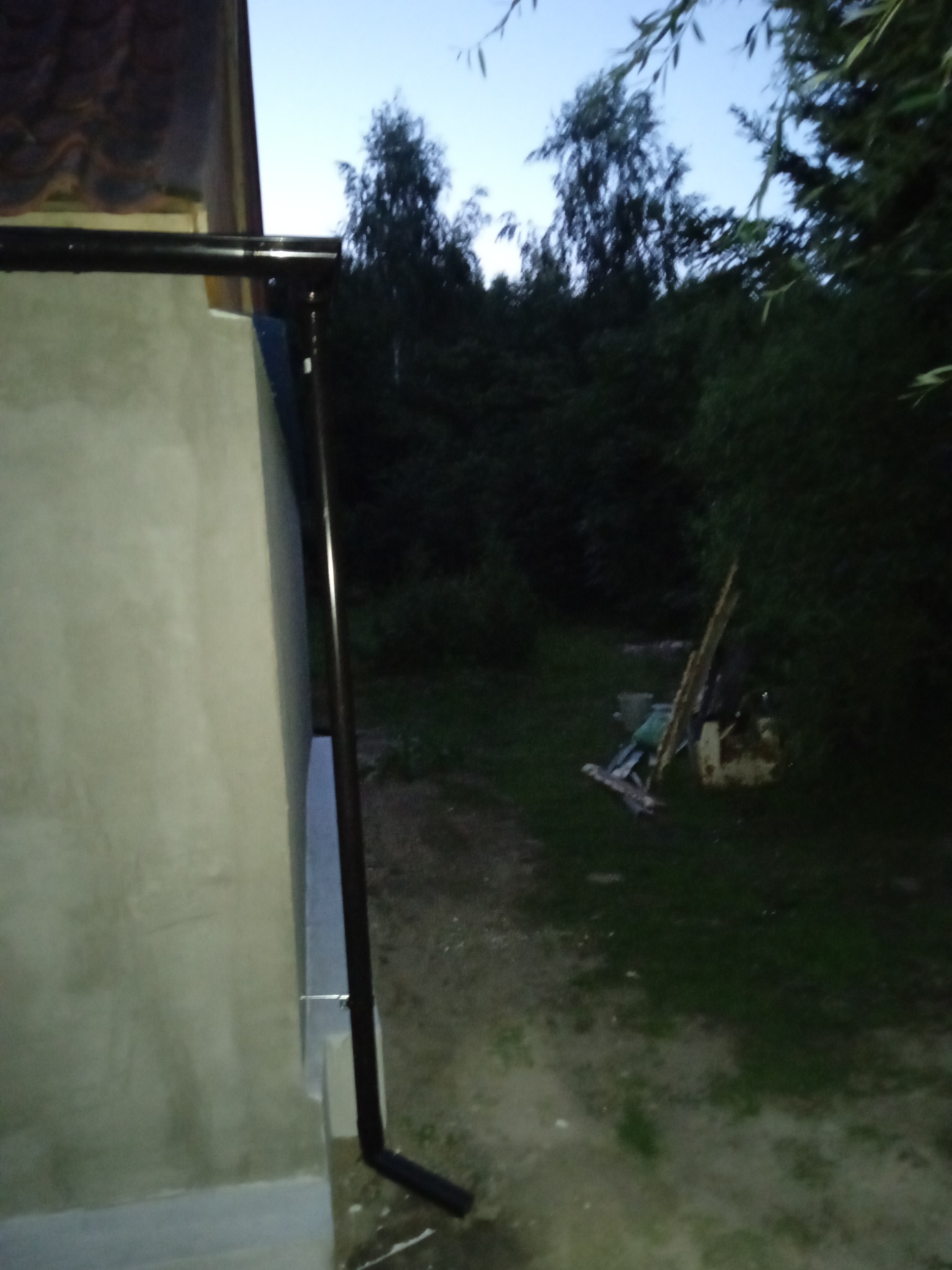 Ściana budynku po tynkowaniu z zamontowaną rynną w ciemnym otoczeniu zieleni.