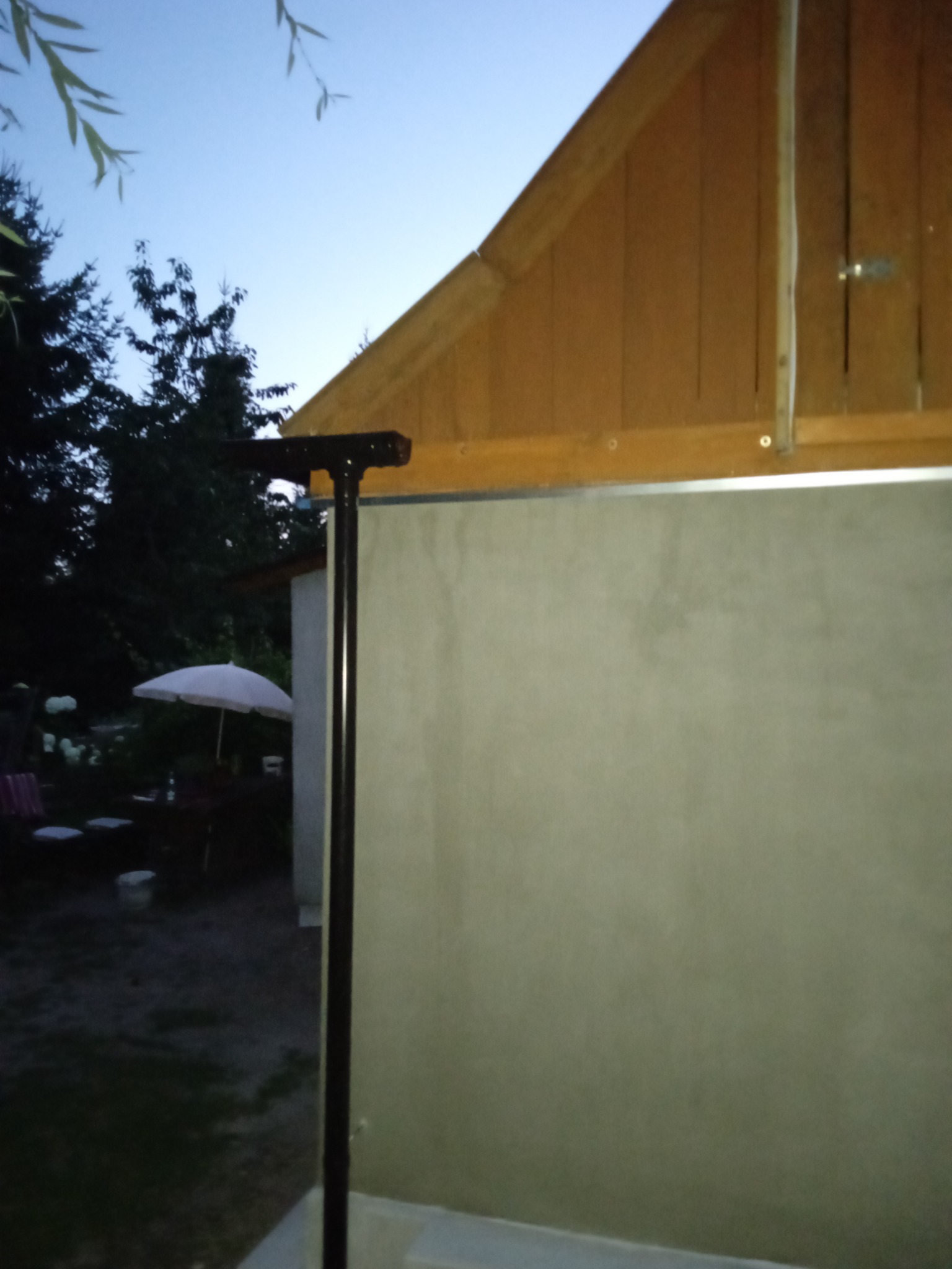 Fragment nowej elewacji budynku w kolorze jasnoszarym, widoczny brązowy dach z drewnianym wykończeniem, ciemna rura spustowa i w tle ogród z parasolem.