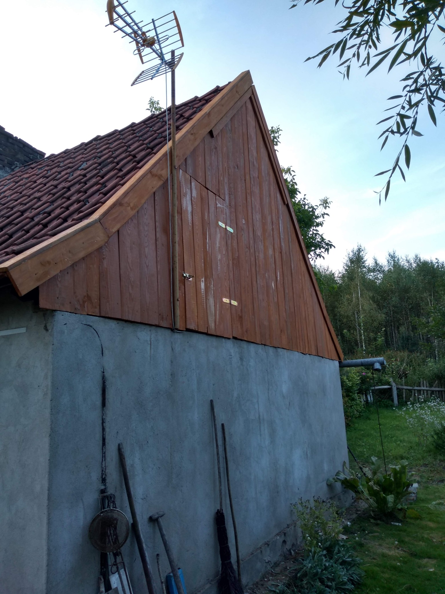 Ściana budynku z nową drewnianą elewacją i anteną telewizyjną na dachu, widoczne narzędzia opierające się o ścianę i zieleń w tle.