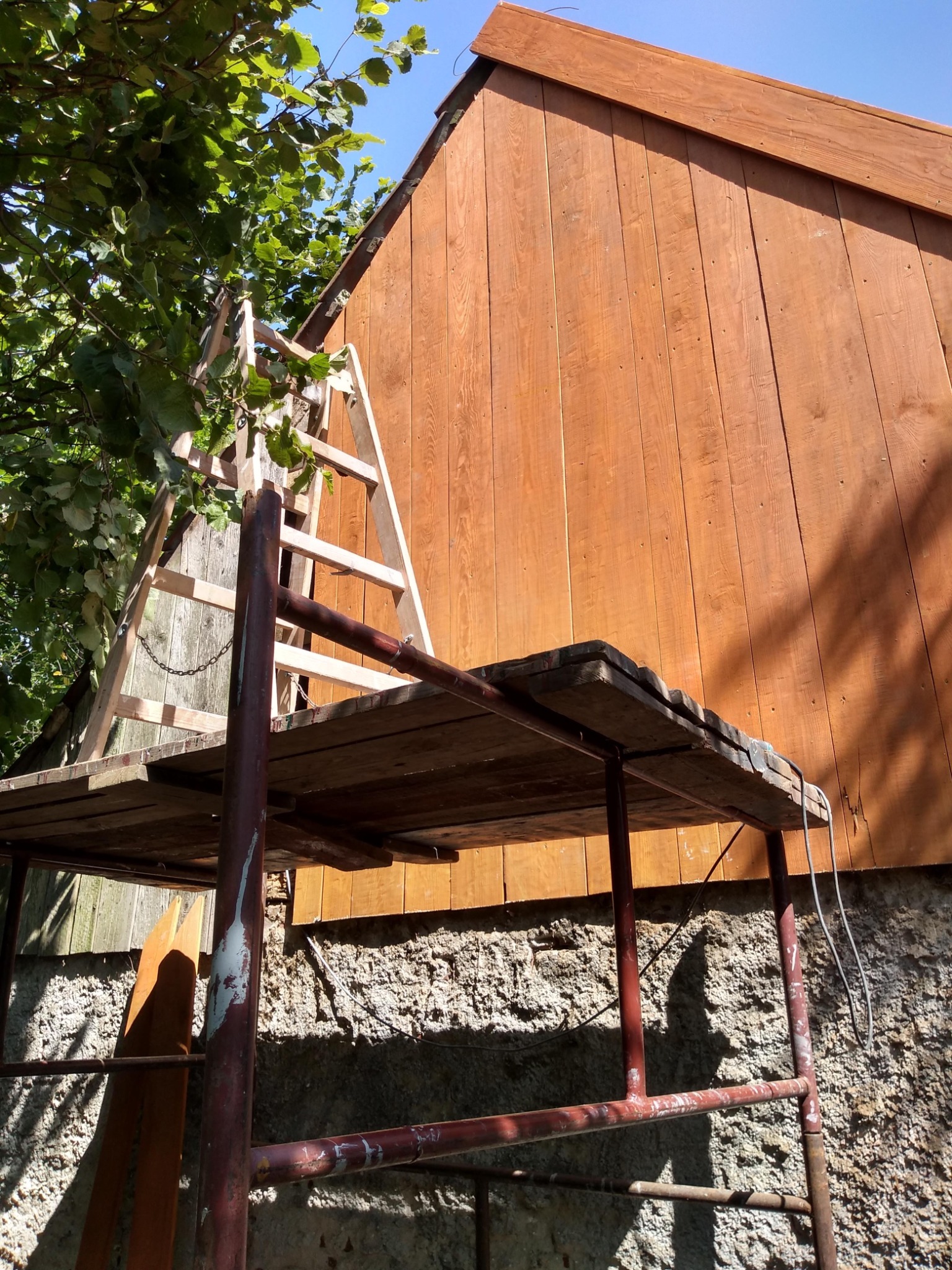 Montaż drewnianej elewacji na budynku, widoczne rusztowanie i drabina oparta o ścianę, częściowo zasłonięte przez gałęzie drzewa.