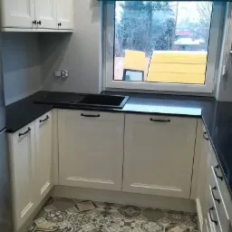 Kuchnia w Białymstoku po remoncie: białe szafki z czarnym blatem i zlewozmywakiem, wzorzysta podłoga z płytek, turkusowa roleta rzymska w oknie z widokiem na żółty samochód dostawczy.