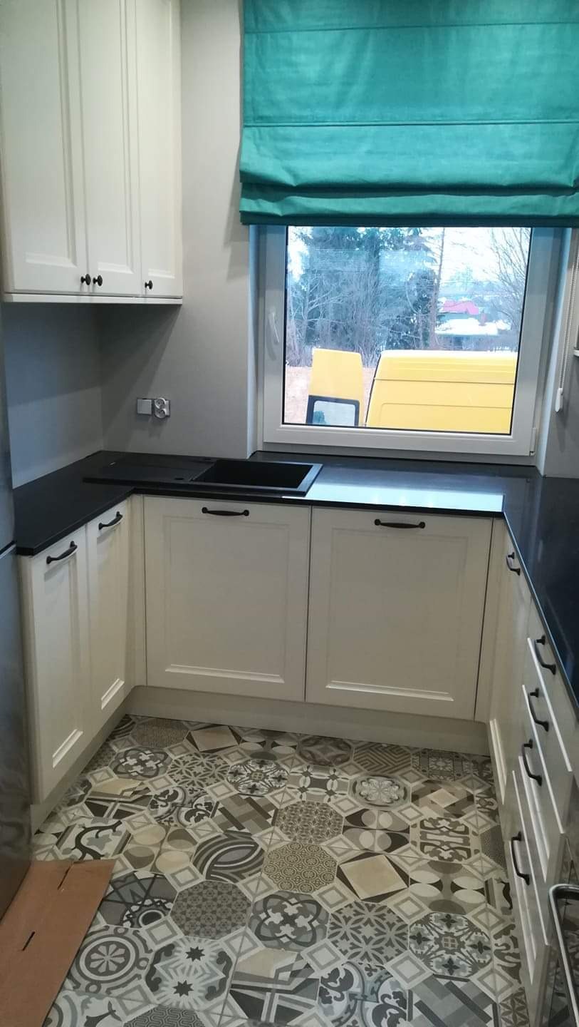 Kuchnia w Białymstoku po remoncie: białe szafki z czarnym blatem i zlewozmywakiem, wzorzysta podłoga z płytek, turkusowa roleta rzymska w oknie z widokiem na żółty samochód dostawczy.