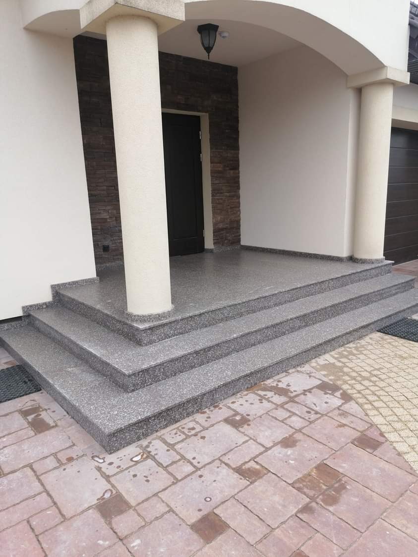 Granitowe schody wejściowe z trzema stopniami, prowadzące do ciemnych drzwi wejściowych, otoczonych kolumnami i kamienną okładziną ściany.