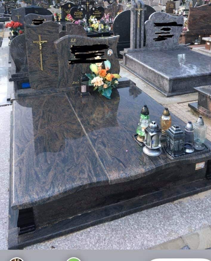 Polerowany nagrobek z ciemnego granitu z wyraźnym wzorem, ozdobiony bukietem kwiatów i lampionami, ustawiony na cmentarzu z widocznymi innymi grobami w tle.