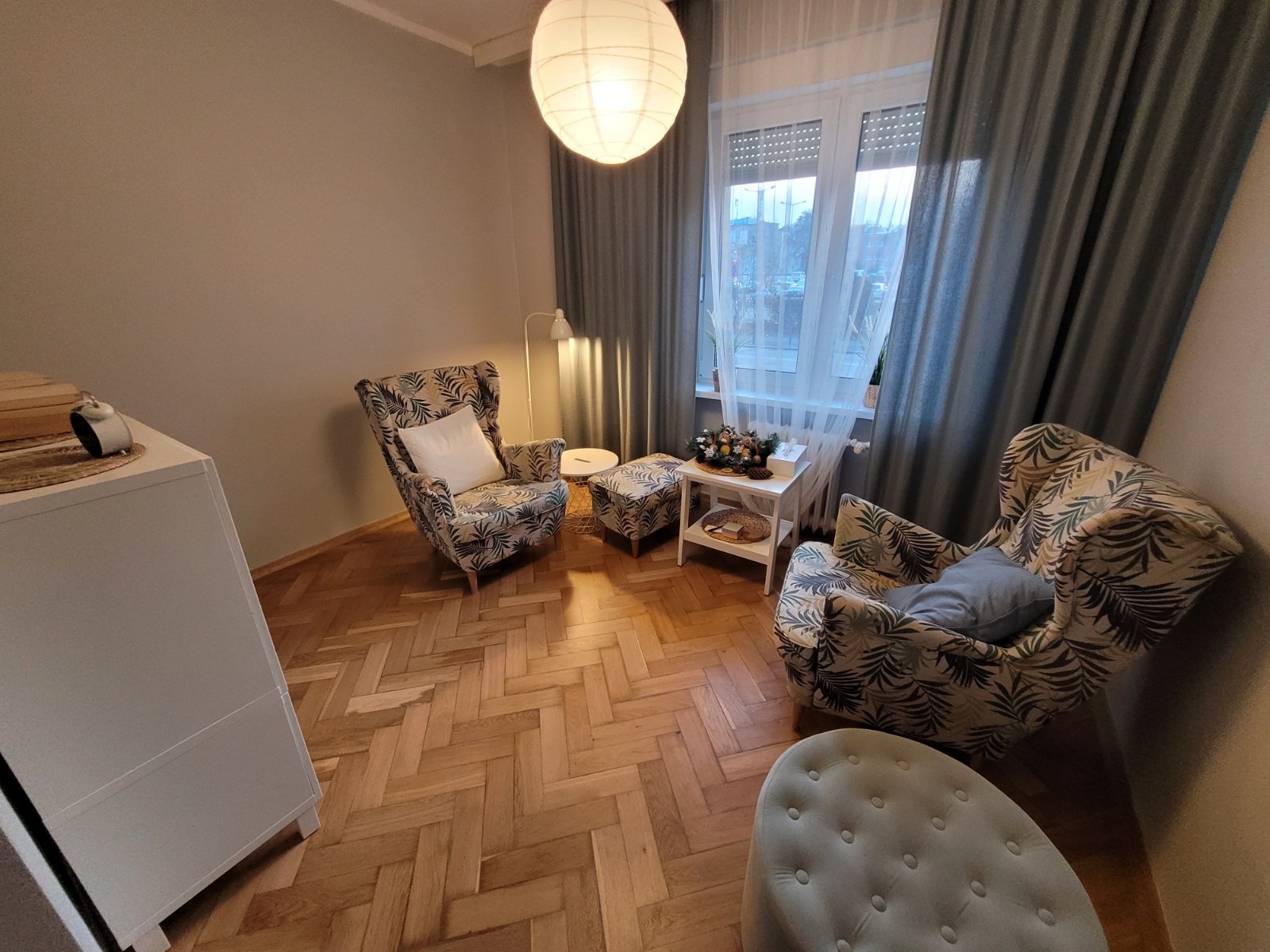 Jasne wnętrze pokoju dziennego z dwoma fotelami w liście, pufami, parkietem w jodełkę i dużym oknem z zasłonami.