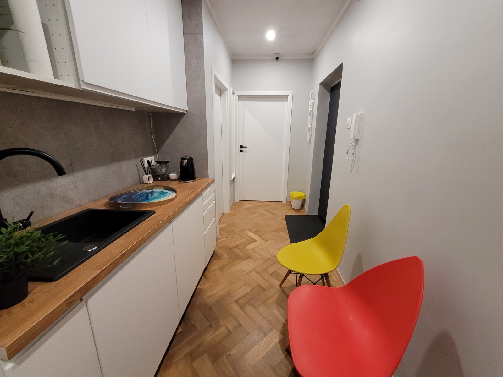 Nowoczesna kuchnia z drewnianym blatem, czarnym zlewem, dwoma krzesłami w stylu retro (czerwonym i żółtym) oraz parkietem ułożonym w jodełkę.