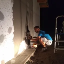 Mężczyzna szpachluje ścianę zewnętrzną przy słabym oświetleniu, widoczny fragment drabiny i wiadro z zaprawą.