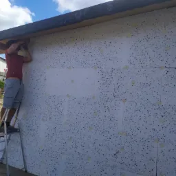 Pracownik na aluminiowej drabinie mocuje białe płyty izolacyjne z czarnymi plamkami do zewnętrznej ściany budynku, używając narzędzia widocznego w rękach, w tle domy i zieleń.