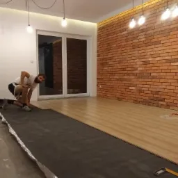 Mężczyzna w trakcie układania paneli podłogowych w pomieszczeniu z ceglaną ścianą i nowoczesnym oświetleniem, widoczna folia izolacyjna i narzędzia.