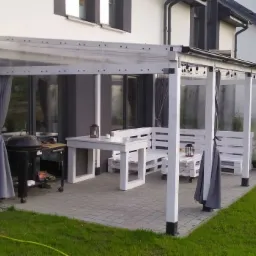 Zadaszona, osłonięta przezroczystą folią i szarymi zasłonami altana z meblami z palet pomalowanymi na biało, grill na kółkach, lampki dekoracyjne, trawnik i kostka brukowa.