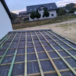 Konstrukcja dachu z drewnianych łat i czarnej membrany hydroizolacyjnej, widok z góry, w tle budynki mieszkalne i drzewa.