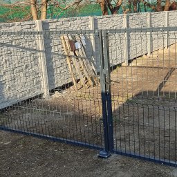 Prywatny - Zamknięta, dwuskrzydłowa brama panelowa w kolorze grafitowym, z betonowymi przęsłami o fakturze kamienia łupanego, widoczna kobieta w tle.