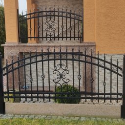 Prywatny - Czarna, metalowa brama wjazdowa i ogrodzenie kute z ozdobnymi elementami, prowadzące do domu z elewacją w odcieniach beżu i brązu.