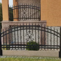 Czarna, metalowa brama wjazdowa i ogrodzenie kute z ozdobnymi elementami, prowadzące do domu z elewacją w odcieniach beżu i brązu.