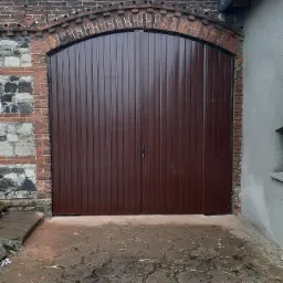 Dwuskrzydłowa, brązowa brama garażowa z pionowymi panelami, osadzona w ceglanym łuku, z widocznymi fragmentami starego muru i brukowanej nawierzchni przed bramą.