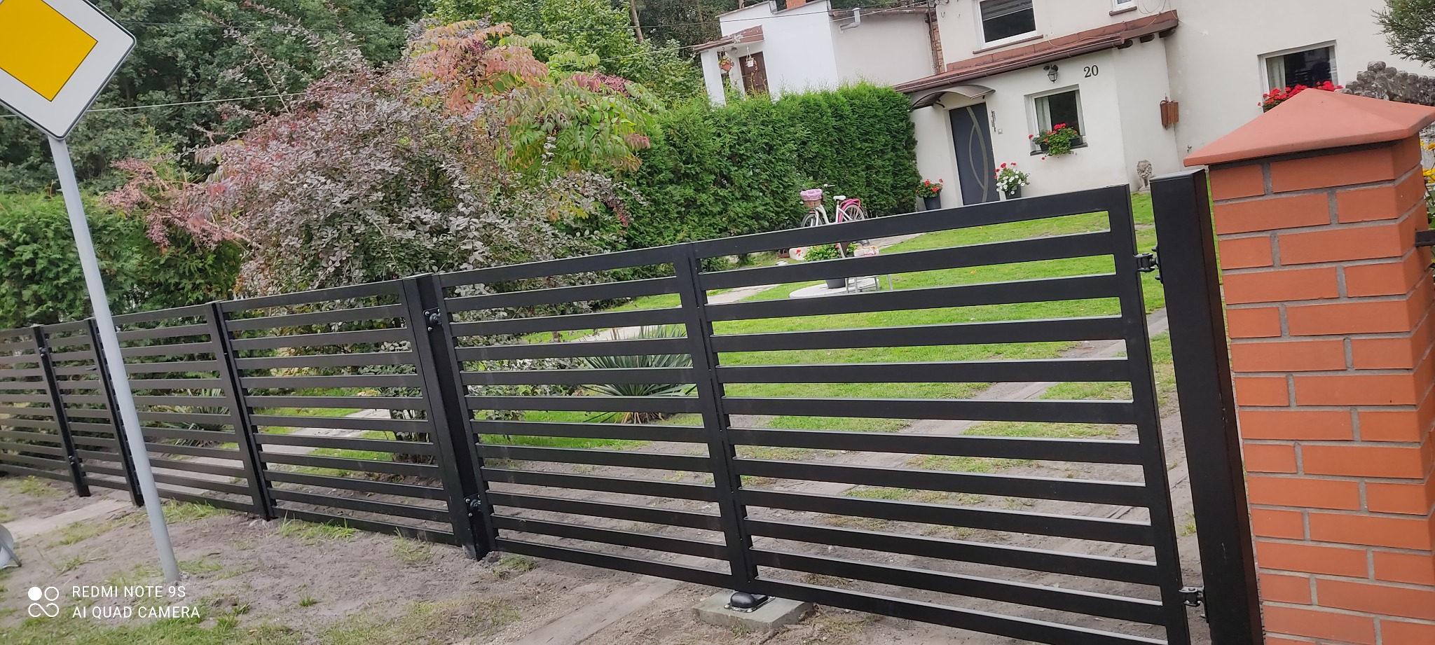 Nowoczesne, poziome, czarne ogrodzenie panelowe z ceglanym słupkiem, na tle zieleni i domu z numerem 20.