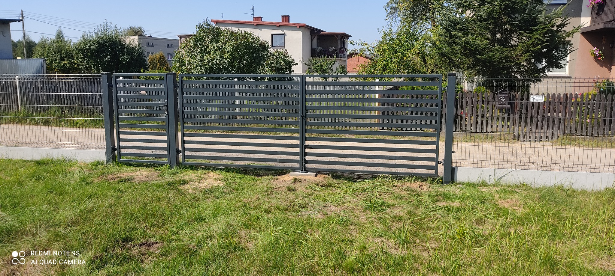 Nowoczesna, pozioma brama wjazdowa z szarych paneli stalowych, widoczna z zewnątrz posesji, z domami i zielenią w tle.