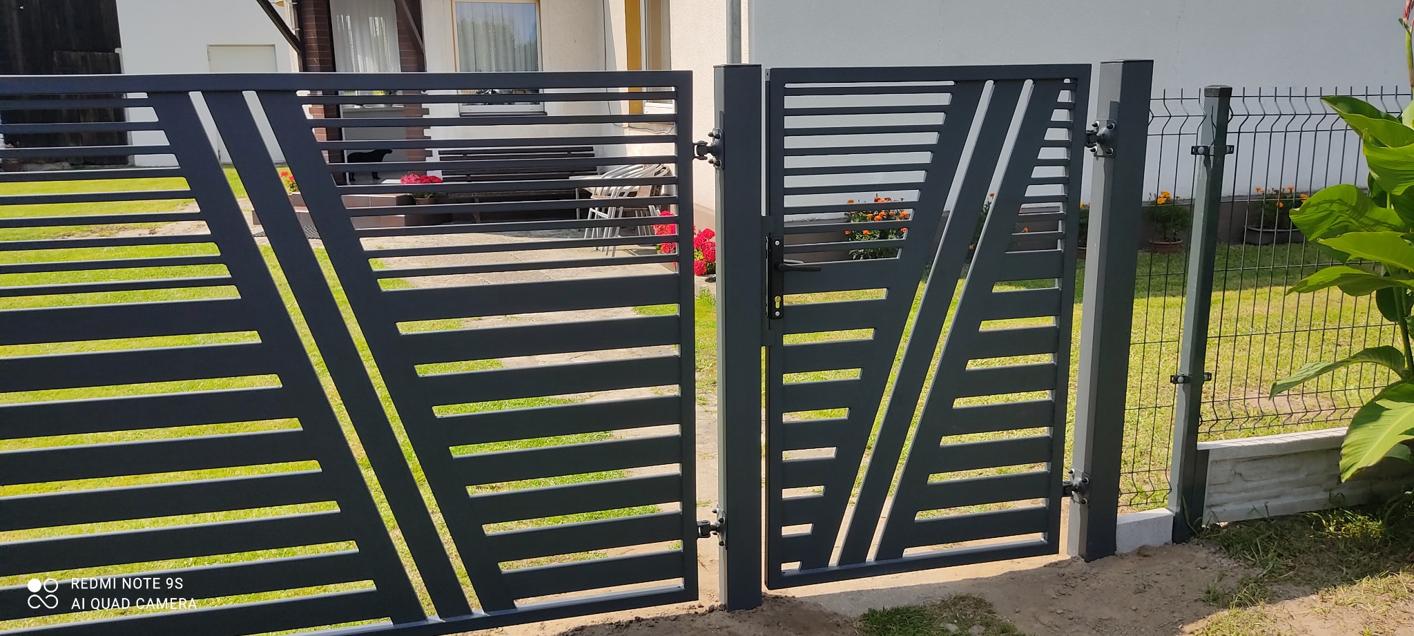 Nowoczesna, grafitowa brama dwuskrzydłowa z poziomymi i ukośnymi szczeblami, widoczna na tle zielonego trawnika i fragmentu domu.