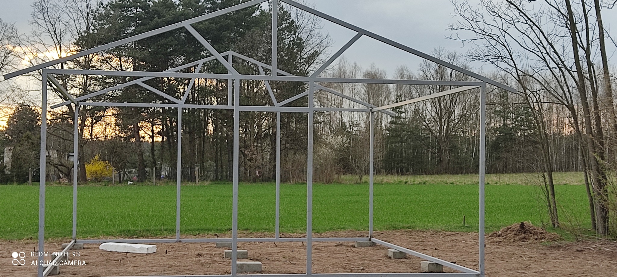 Metalowa konstrukcja hali w trakcie budowy, widoczne fundamenty z bloczków betonowych, tło stanowi zielone pole i drzewa, pochmurne niebo.