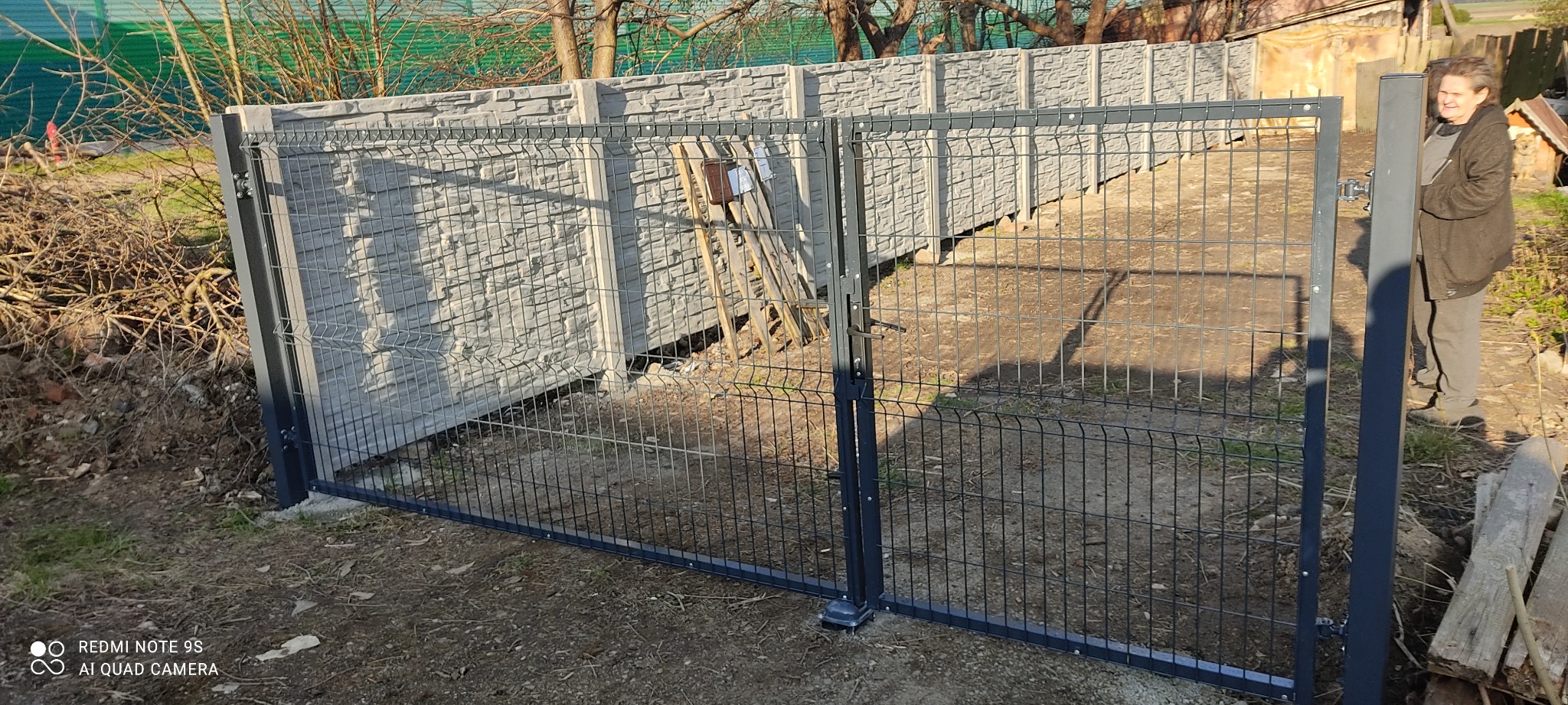 Zamknięta, dwuskrzydłowa brama panelowa w kolorze grafitowym, z betonowymi przęsłami o fakturze kamienia łupanego, widoczna kobieta w tle.