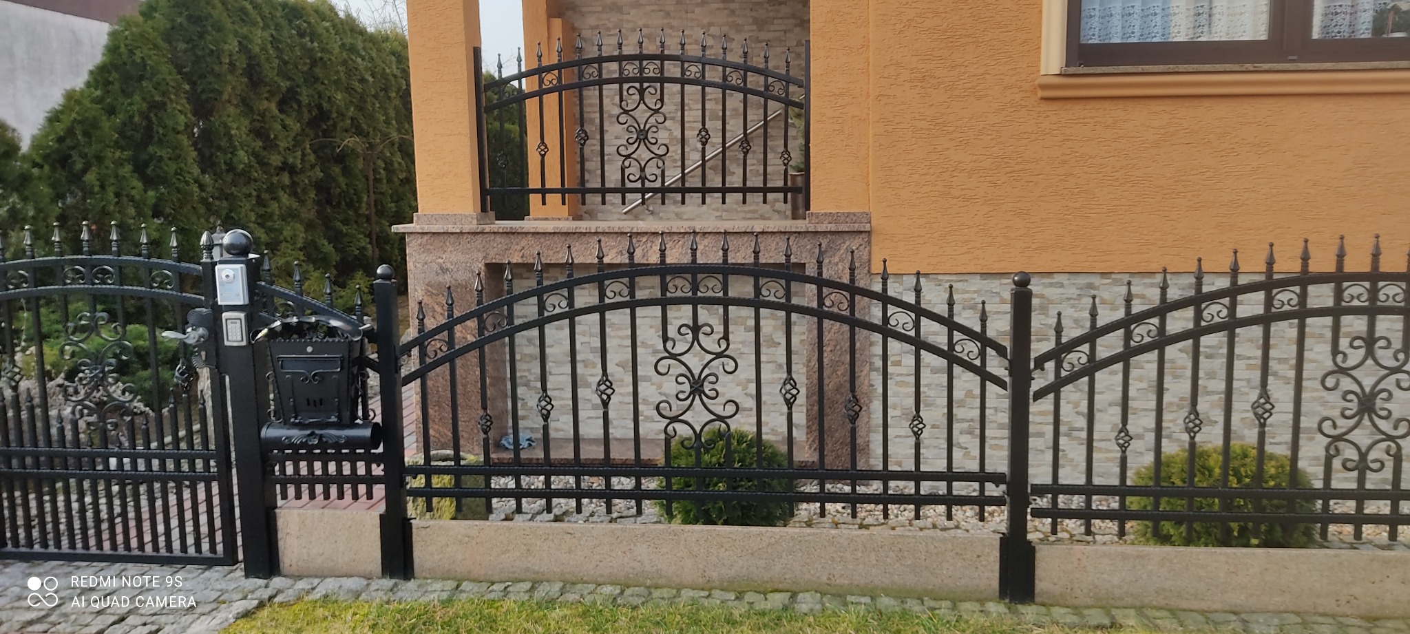 Czarna, metalowa brama wjazdowa i ogrodzenie kute z ozdobnymi elementami, prowadzące do domu z elewacją w odcieniach beżu i brązu.