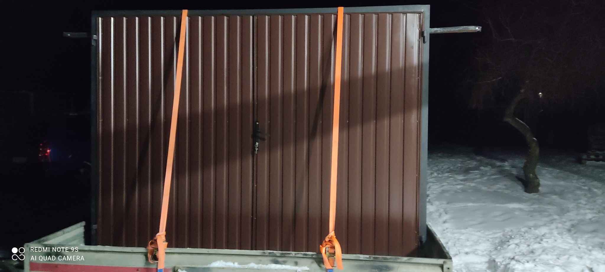 Brązowa dwuskrzydłowa brama garażowa z blachy trapezowej, zabezpieczona pasami transportowymi na przyczepie w zimowej scenerii.